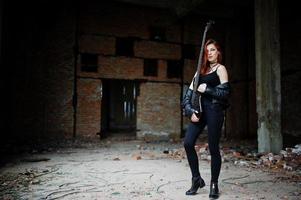 Red haired punk girl wear on black with bass guitar at abadoned place. Portrait of gothic woman musician. photo