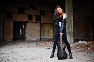 Red haired punk girl wear on black with bass guitar at abadoned place. Portrait of gothic woman musician. photo