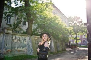 mujer rubia con vestido negro, chaqueta de cuero, collares y sombrero posado contra la calle de la ciudad al atardecer. foto