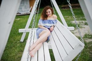 Portrait of a stunning young girl wearing blue marine-styled dress sitting on swings on the lakeside. photo