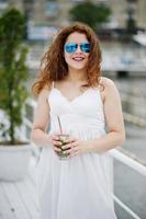 retrato de una joven atractiva posando con su cóctel en un lago con gafas de sol. foto