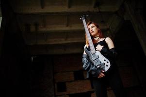 Red haired punk girl wear on black with bass guitar at abadoned place. Portrait of gothic woman musician. photo