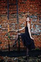 Red haired punk girl wear on black dress at the roof against brick wall with iron ladder. photo