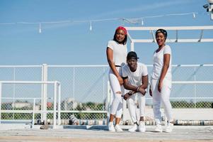 Three stylish african american friends, wear on white clothes at pier on beach. Street fashion of young black people. Black man with two african girls. photo