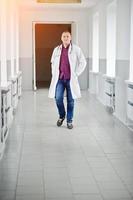 Handsome and confident young doctor in white coat with stethoscope posing in the hospital. photo