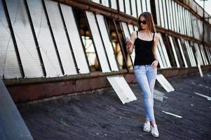 chica con gafas de sol y jeans, con teléfono y auriculares posados en el techo de un lugar industrial abandonado con ventanas. foto