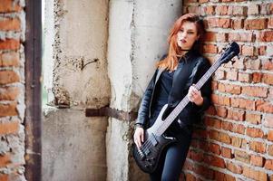 Red haired punk girl wear on black with bass guitar at abadoned place. Portrait of gothic woman musician. photo