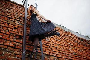 Chica punk de pelo rojo vestida de negro en el techo contra una pared de ladrillo con escalera de hierro. foto