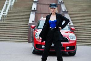 Portrait of beautiful blonde sexy fashion woman model in cap and in all black, blue corset, with bright makeup near red city car. photo