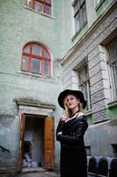 Blonde woman on black dress, leather jacket, necklaces and hat against old street of city. photo