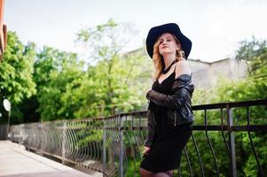 mujer rubia con vestido negro, chaqueta de cuero, collares y sombrero contra barandillas. foto