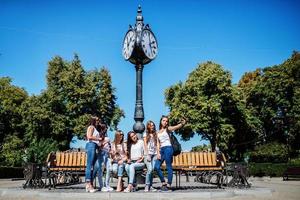 Six beautiful young girls sitting on a bench next to the old street clock in the park. photo