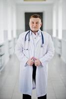 Handsome and confident young doctor in white coat with stethoscope posing in the hospital. photo