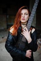 Red haired punk girl wear on black with bass guitar at abadoned place. Portrait of gothic woman musician. photo