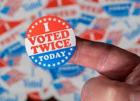 Finger with I Voted Twice sticker in front of many election voting badges to illustrate voter fraud photo