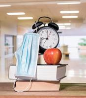 Back to school background concept with stack of books, alarm clock, apple with face mask photo