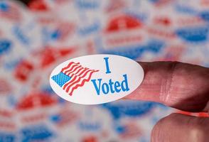 Finger with I Voted sticker in front of background created from many election voting badges photo