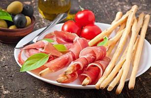 Grissini bread sticks with ham, olives, basil on old wooden background photo