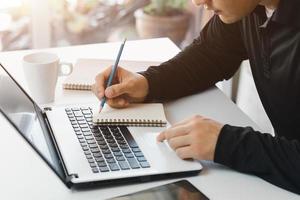 young man using laptop computer and mobile phone When looking for financial information in business, work at the desk. Writing with a pen, studying remotely from home and working from home. photo