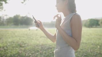 ung vuxen arbetande kvinna som använder smartphone som går ensam under träd i utomhusparken, mänsklig naturteknik, sidovy dam kommer online via mobiltelefon, skicka svarsmail, soligt dagsljus video