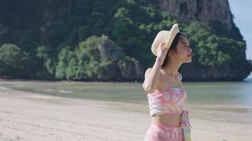 Young pretty asian female walking along Island alone with wind blowing hairs and hat, Travel summer vacation, Asian lady wear straw hat Enjoy summer Holiday, tropical island season hot sunny weather video