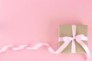 Brown paper gift box with pink satin curly ribbon bow on pastel pink background. Flat lay mother day, father day, valentines day, birthday concepts with copy space.