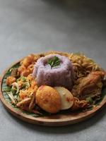 High angle view of purple rice with Indonesian traditional condiments, like spicy egg, fried noodles, spicy chicken, fried potato, on wooden plate and uneven grey color table.