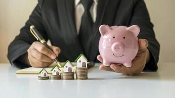Businessman holding piggy bank in hand and house model on the pile of coins, the concept of saving money and planning to buy a house.