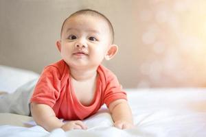 familia feliz, lindo bebé recién nacido asiático usa camisa roja acostado, gateando, juega en una cama blanca con una sonrisa sonriente cara feliz. pequeño inocente niño adorable niño en el primer día de vida. concepto del día de la madre. foto