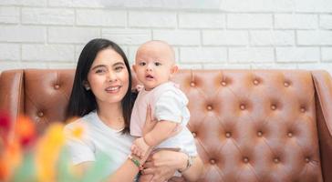 Happy family, Cute Asian newborn baby with young mother smile happy face sitting sofa at home. Innocent little infant adorable.  Mom carrying, taking care with love. Parenthood, mother day concept. photo