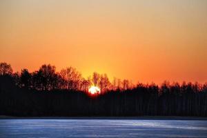 Sun setting behind black forest silhouette over frozen lake iced surface photo