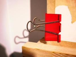 Red big paper clip holding a paper sheet corner on wood with strong light causing a deep shadow on wall photo