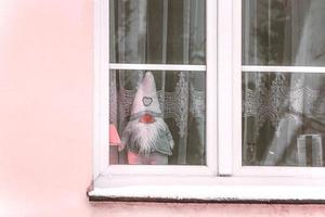 gnomo de juguete de fieltro parado detrás de la ventana con cortina de tul blanco en un edificio de pared rosa foto