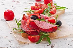 Sándwich con salchicha, aceitunas, tomate y rúcula en el fondo de madera antiguo foto