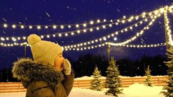 A girl looks at flying snowflakes on a winter evening and eats snow from mittens against the background of fairy lights of outdoor garlands. Christmas, New Year mood. Slow motion video