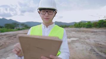 asiatischer männlicher ingenieur trägt sicherheitswesten-schutzhelm, der digitales tablet auf outdoor-site betrachtet, fachwissen, bauplanungsfortschritt, outdoor-dreckland, hart arbeitender mann video