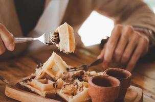 Belgian waffles with chocolate, berries and powdered sugar, a hand with a knife and fork, glass cup with coffee in the process of eating. photo