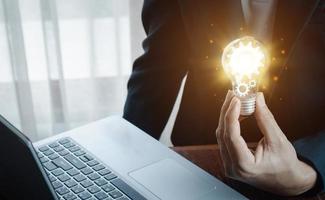 The hand of a man with a light bulb And there is a gear icon in it And he is using a notebook computer. photo