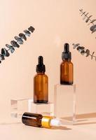 Glass dropper bottles with a pippette with black rubber tip on glass podium with eucalyptus leaves and beige background, mockup design photo
