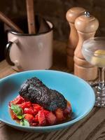 lomo de cerdo al horno marinado en tinta de sepia con tomates rojos y albahaca en plato azul, sal, pimienta, cubiertos y cóctel de alcohol de vermú seco con oliva en la mesa del bar