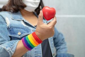 Asian lady wearing rainbow flag wristbands and hold red heart, symbol of LGBT pride month celebrate annual in June social of gay, lesbian, bisexual, transgender, human rights. photo