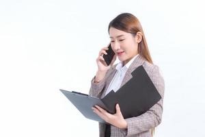 una trabajadora asiática con traje formal y camisa blanca llama por teléfono y abre un archivo de documento o un portapapeles para verificar los datos.