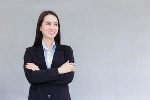 una mujer asiática que trabaja en un negocio profesional que usa un traje formal negro se para con el brazo cruzado y sonríe alegremente en la oficina como fondo. foto