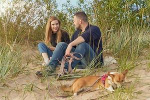 joven pareja feliz con perro corgi, hombre y mujer sentados en la arena. dos personas en ropa de abrigo foto