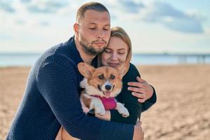 Young happy couple with dog standing on beach. Beautiful girl and guy and Corgi puppy having fun photo