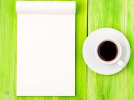 bloc de notas abierto con una página en blanco para escribir una idea o una lista de tareas pendientes, una taza de café en una mesa de madera verde brillante foto