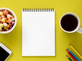 Open notepad on spiral with clean white page, cup with tea, caramels in a bowl, smartphone and color pencil on bright yellow background color photo
