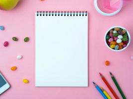 Open notebook with a clean white sheet, sweets, mobile phone, crayon, decorations on a pink bright table photo