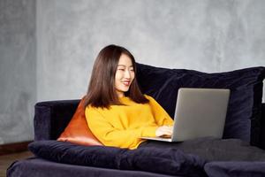 mujer asiática mirando la computadora portátil, trabajando en casa, acostada en el sofá. hermosa mujer japonesa foto