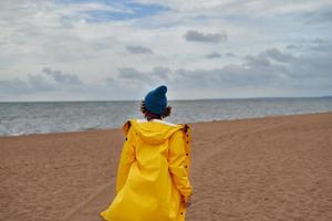 Back view of woman in bright yellow cloak and red hat on shore of North sea on autumn day